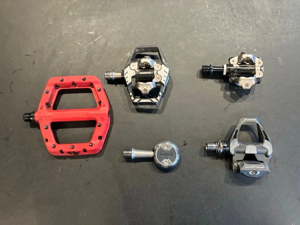 Bicycle pedals of various types, including clipless and flat designs, displayed on a dark surface.