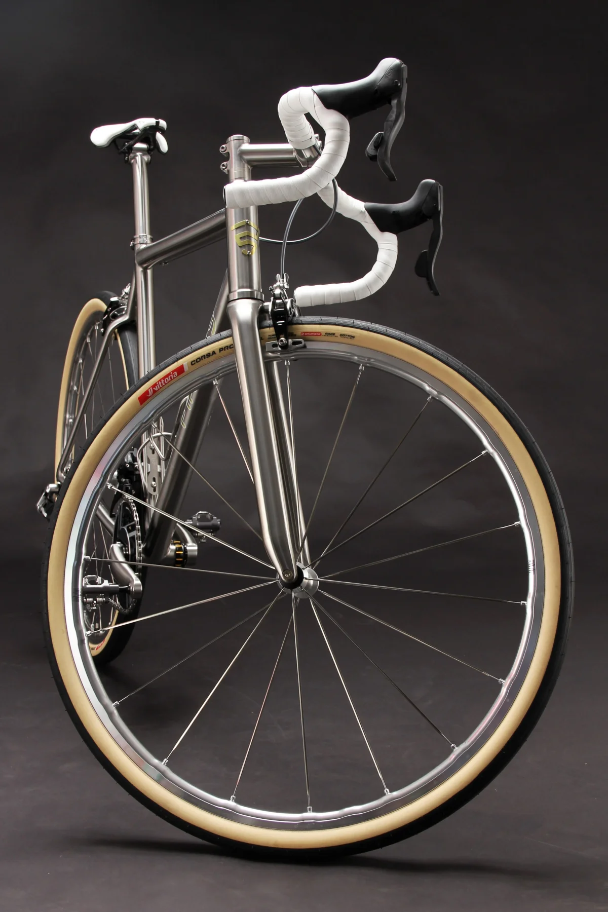 A sleek silver bicycle with drop handlebars and beige tires against a dark background. A sleek silver bicycle with drop handlebars and beige tires against a dark background.